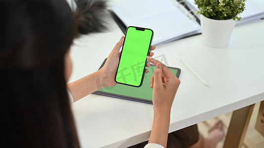 裁剪拍摄的女人拿着智能手机与办公桌上的绿色铬屏幕。