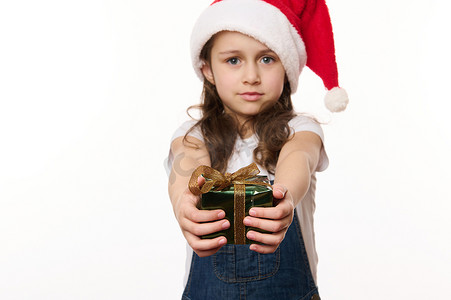 小孩圣诞老人摄影照片_可爱的女婴，一个戴着圣诞帽和牛仔服的小孩，递着一份快乐的圣诞礼物，看着镜头
