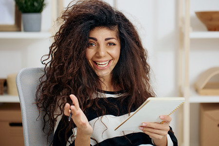 穿着条纹衬衫的快乐微笑快乐漂亮卷发晒黑的拉丁女人拿着笔记本看着相机说是的在家庭内部写工作信。