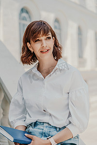 一个身穿白衬衫和蓝色牛仔裙的女人坐在矮墙上，在建筑物的背景下，手里拿着一本蓝色的笔记本。