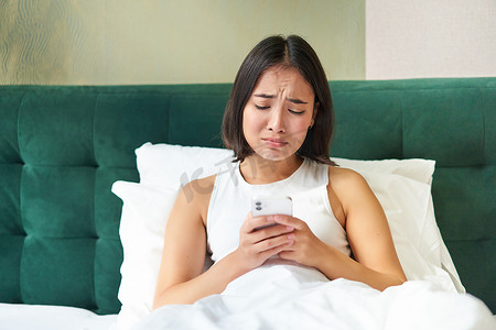 失望的亚洲女孩，哭泣和抱怨，心烦意乱，躺在床上枕头上，拿着手机，读着令人沮丧的坏消息，在糟糕的心情中醒来