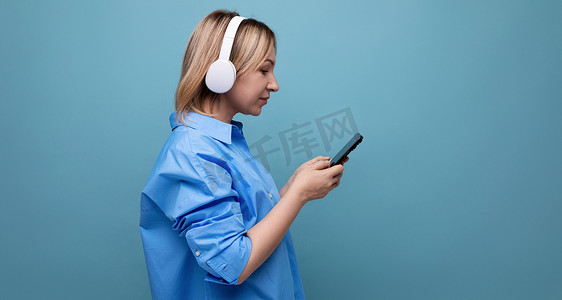 一位细心的千禧一代女性穿着休闲衬衫、戴着白色大耳机、手里拿着智能手机的横向照片，背景是蓝色，有复制空间