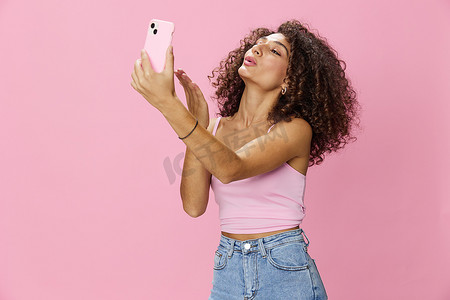 女博主手持电话视频通话，卷发粉色上衣，牛仔裤摆在粉色背景、复制空间、技术和社交媒体上，在线