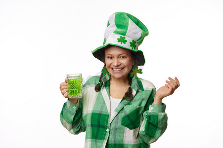 戴着带三叶草叶子的嘉年华帽的可爱女人，对着镜头微笑，拿着一杯绿色啤酒，庆祝圣帕特里克节