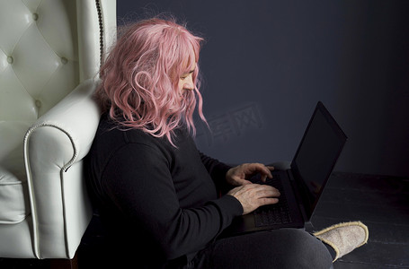 假发摄影照片_一个戴着粉色假发的男人坐在白色椅子附近的地板上，在电脑前工作
