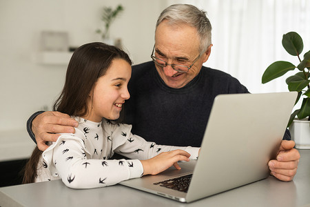 快乐的退休祖父和漂亮的孙女一边笑一边看电子书，通过笔记本电脑一起学习教育。