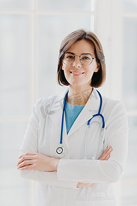 美丽开朗的女医生穿着白色长袍、眼镜和语音内窥镜，双手交叉，自信地看着镜头，留着黑色短发，站在白色背景下，在诊所工作