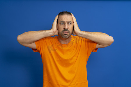 身穿橙色 T 恤、双手托着头的拉丁男子惊呆了，与蓝色工作室背景隔离开来。