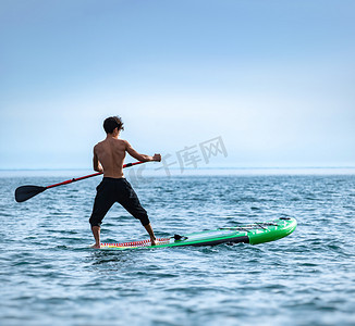白天，一个运动型男在海上划着桨在冲浪板上游泳，在美丽的天空下