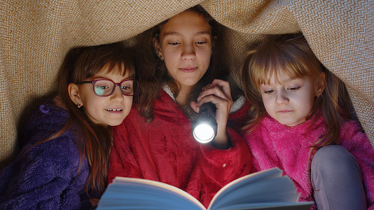 孩子们在毯子下在家里用手电筒阅读有趣的童话故事书