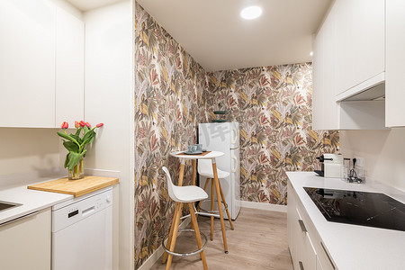 壁柜摄影照片_紧凑型厨房配有小桌子和舒适的烹饪台面，配有现代电器和带花卉壁纸的壁柜。