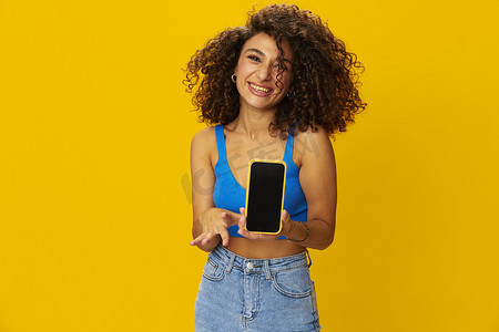 手持电话的有影响力的女性在视频通话中自拍自由职业者社交媒体技术工作，蓝色衬衫黄色背景的卷发爆炸头，牙齿微笑，复制空间