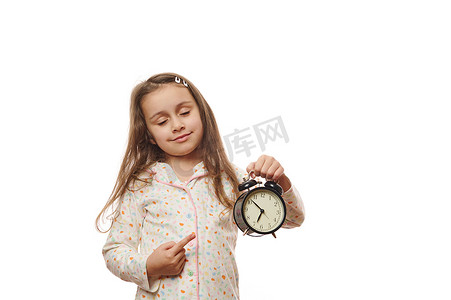 睡着的睡着的小女孩穿着白色睡衣，闭着眼睛，指着显示就寝时间的闹钟