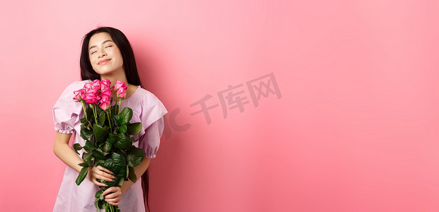 梦幻般的亚洲少女感觉浪漫，手捧鲜花做梦，想象情人节约会，穿着可爱的裙子，站在粉色背景上