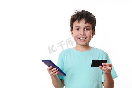 移动电子支付摄影照片_迷人的青春期男孩，穿着蓝色 T 恤、带智能手机和塑料信用卡的学童，与世隔绝在白色背景中