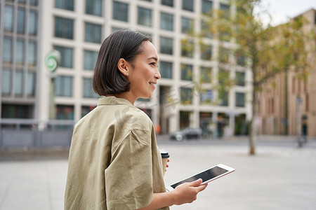 时尚都市女孩的肖像带着平板电脑在城市里散步，喝着外卖咖啡