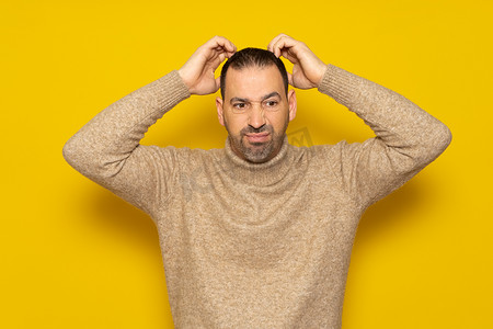 米色摄影照片_留着胡子的西班牙裔男子身穿米色高领毛衣，在试图解决问题或疑问时精力充沛地挠头，被黄色背景隔离。