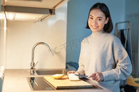 三明治背景摄影照片_微笑的亚洲女人做三明治，在厨房里切面包，看着相机