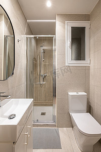现代紧凑型米色瓷砖浴室配有玻璃步入式淋浴间、水槽和镜子以及带窗户的白色卫生间。