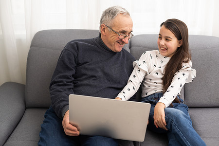 看电子书摄影照片_快乐的退休祖父和漂亮的孙女一边笑一边看电子书，通过笔记本电脑一起学习教育。