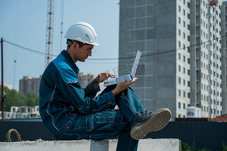 戴安全帽的白人男性建筑工人坐在楼板上，在建筑工地使用笔记本电脑。