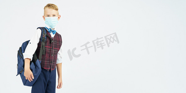 一个脸上戴着医用口罩的小学生背着书包，侧身站立，看着镜头。