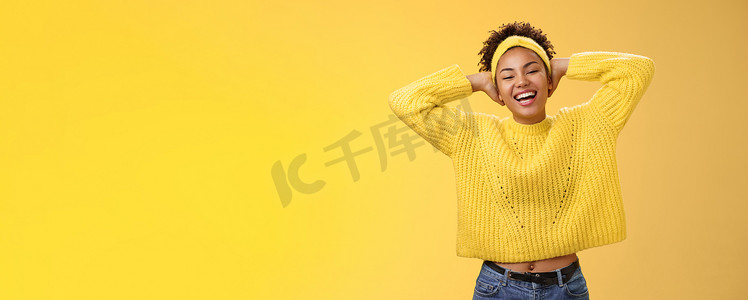 穿着毛衣头带的无忧无虑、快乐幸运的非洲裔美国女学生把手放在脑后，放松地摆出令人不寒而栗的姿势，周末放假，笑得大大的，黄色背景