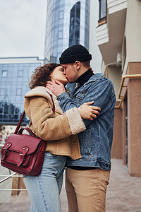 保暖衣服摄影照片_在商业大楼附近的城市里，一对穿着休闲保暖衣服的快乐情侣在户外接吻