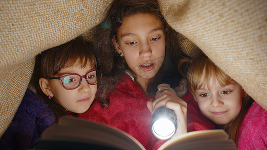 故事童话故事摄影照片_姐妹们在家里用手电筒在毯子下阅读有趣的童话故事书