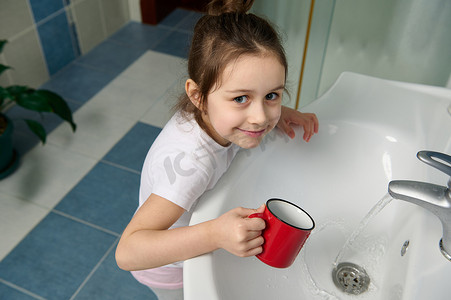 淘气摄影照片_淘气的女婴站在家庭浴室的水槽旁，微笑着看着镜头，同时清洁嘴巴、刷牙