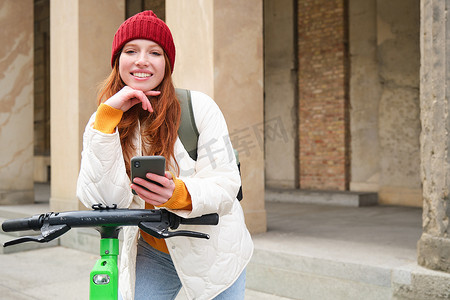 带着手机微笑的年轻红发女性，使用智能手机应用租用电动滑板车，在城市、城市交通和人的概念中骑行