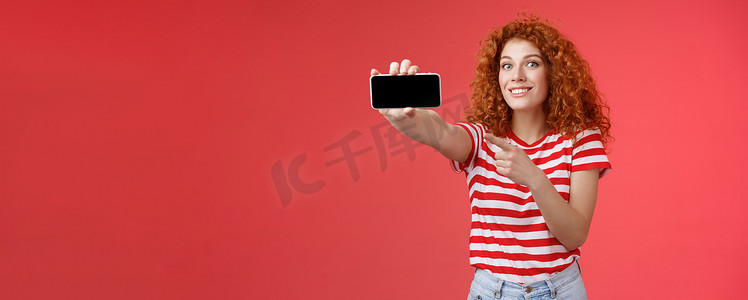 兴奋快乐好看的红发卷发女性展示水平智能手机屏幕指向显示小工具微笑高兴自豪击败朋友得分游戏站立红色背景