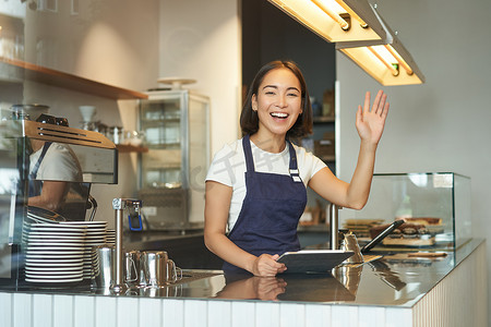 快乐的亚洲女咖啡师，请说下一个，举起手臂邀请咖啡馆顾客点咖啡，站在柜台后面，拿着 POS 终端平板电脑