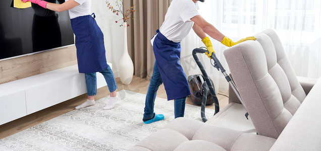 家政团队摄影照片_穿着蓝色制服的专业清洁工洗地板，擦去公寓客厅家具上的灰尘。