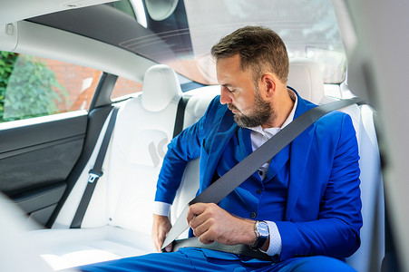 一个身穿蓝色西装的白人男子在汽车后座上系好安全带。