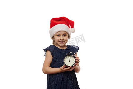 戴着圣诞帽的快乐小女孩在相机的表盘上展示午夜的闹钟，为即将到来的冬季活动欢欣鼓舞