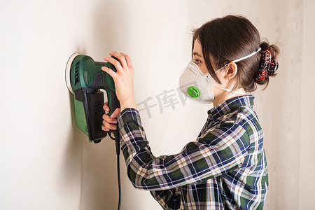 戴防护面罩的妇女用电动砂光机打磨石膏墙面
