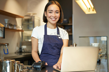 微笑的亚洲女咖啡师，在咖啡馆工作，为客户服务，一边看笔记本电脑，一边使用读卡器收款，卖咖啡