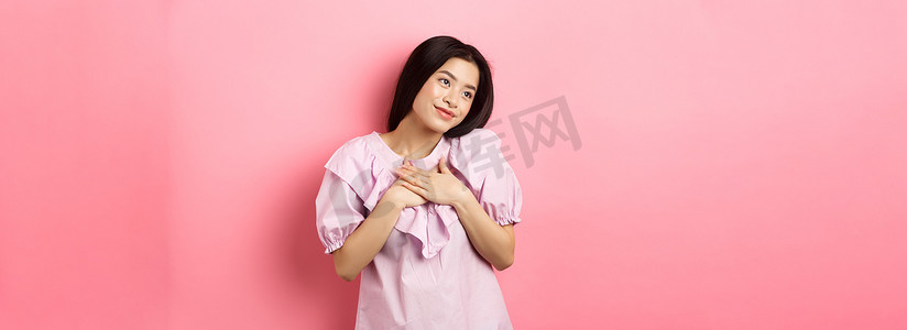 梦幻般的亚洲女孩微笑着，手放在胸前，向左看一些浪漫可爱的东西，站在粉色背景上
