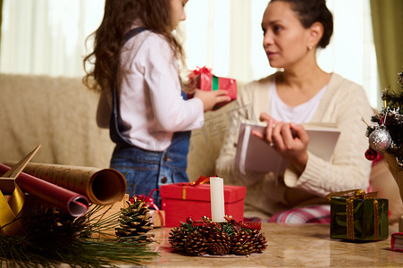专注于蜡烛、包装材料和松果，背景模糊的可爱女婴给妈妈送圣诞礼物
