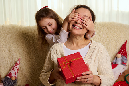 慈爱的母亲微笑着，可爱的女儿用手遮住眼睛，用时尚的红盒送圣诞礼物