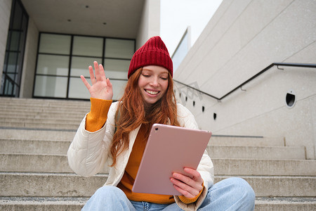 快乐的红发少女，带数字平板电脑的学生，打招呼，对着小工具相机挥手，连接到视频聊天，在应用程序中与朋友交谈