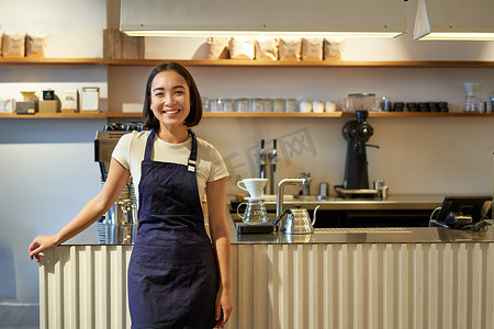 微笑的亚洲女咖啡师的画像，穿着围裙，站在柜台附近喝咖啡，在咖啡馆工作