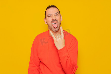 摇滚手势摄影照片_留着胡须的西班牙裔男子身穿红色套头衫，与黄色背景隔离，用手指摆出摇滚手势，伸出舌头。