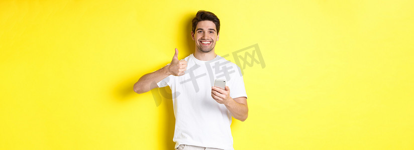 广告黄色摄影照片_快乐满意的男人拿着智能手机，竖起大拇指表示赞同，在网上推荐一些东西，站在黄色背景上