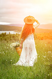 背头摄影照片_一位身穿白色连衣裙、头戴棕色帽子的中年妇女背靠绿色田野站立，手里拿着一个篮子，篮子里放着一大束雏菊。