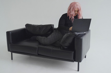 一个戴着粉色假发的男人坐在沙发上，在笔记本电脑上打字的肖像。