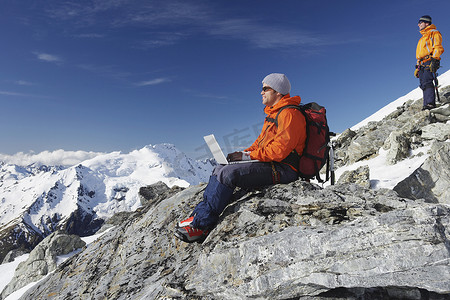 站在山峰上摄影照片_在山峰上使用笔记本电脑的男性登山者的侧视图