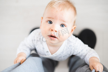 运动裤摄影照片_从上到下看快乐的男婴在家里抱着父亲的运动裤迈出第一步。