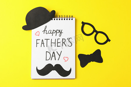 创意父亲节快乐摄影照片_笔记本上刻有父亲节快乐、装饰性领结、眼镜、胡须和彩色背景上的帽子、文字空间
